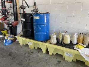 Two large drums—one black and one blue—each fitted with a pump, stand on a yellow spill containment platform. To the right, plastic jugs with long pouring spouts containing various garage fluids, rest on a second spill containment platform. Pipes, cleaning supplies, and equipment are visible in the surrounding industrial workspace.